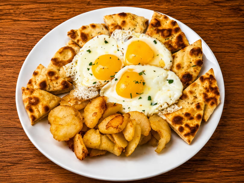 A white plate with three fried eggs, roasted potatoes, and pita bread on a wooden table.