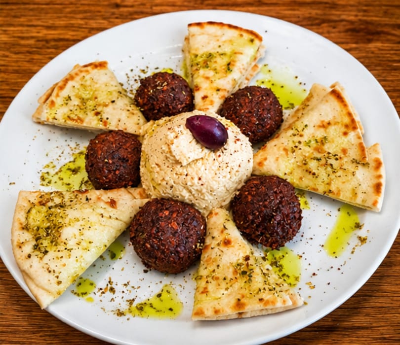 A white plate with hummus, falafel, pita bread, olive, and olive oil drizzle.