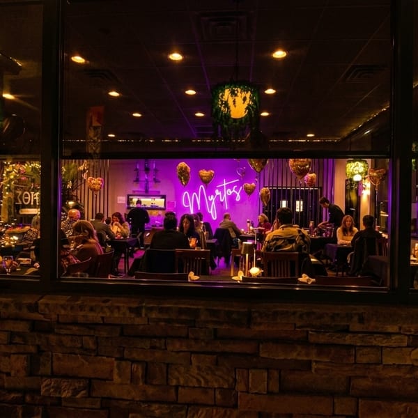 Diners enjoy a lively evening inside Myrtos restaurant, visible through large windows from a brick exterior.