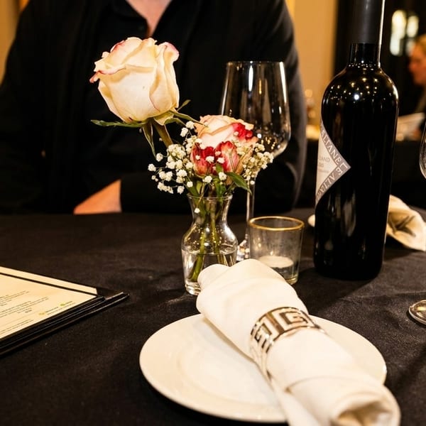 A restaurant table set with a menu, wine bottle, wine glass, and a small vase of roses.